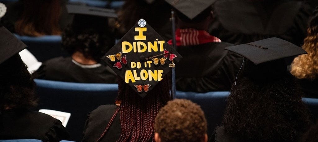 A photograph of Tufts graduates at commencement. The image features the back of new graduate's head's, so their caps are visible. In the middle one cap stands out, which is decorated to read "I Didn't Do It Alone" in big yellow letters with butterflies for decoration.